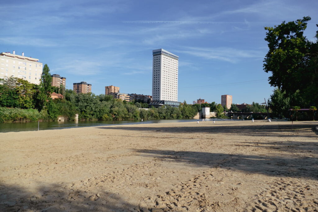 Zona de arena junto al río Pisuerga en la Playa de las Moreras de Valladolid con edificios al fondo